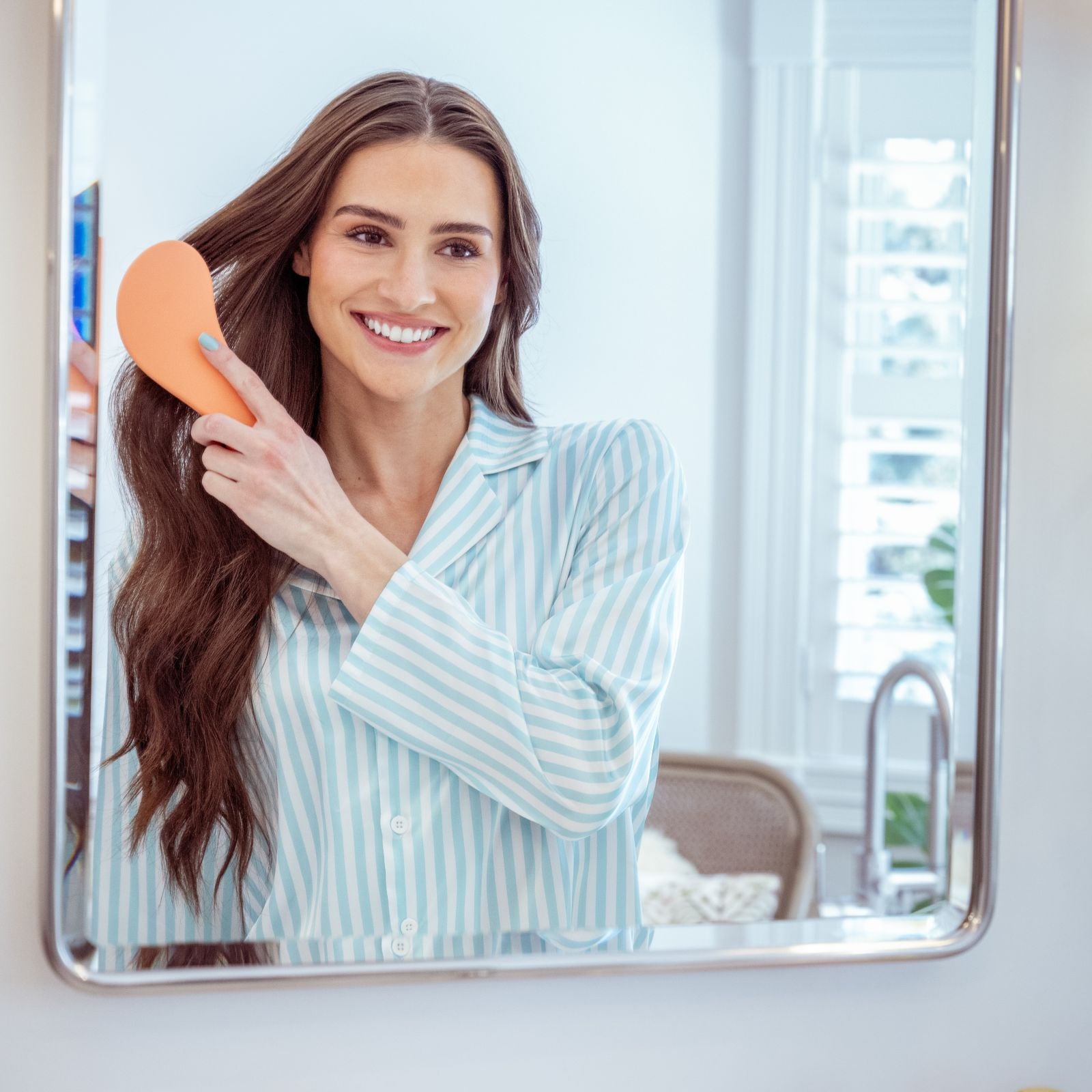 Woman brushing her hair in front of a mirror in a bright room.