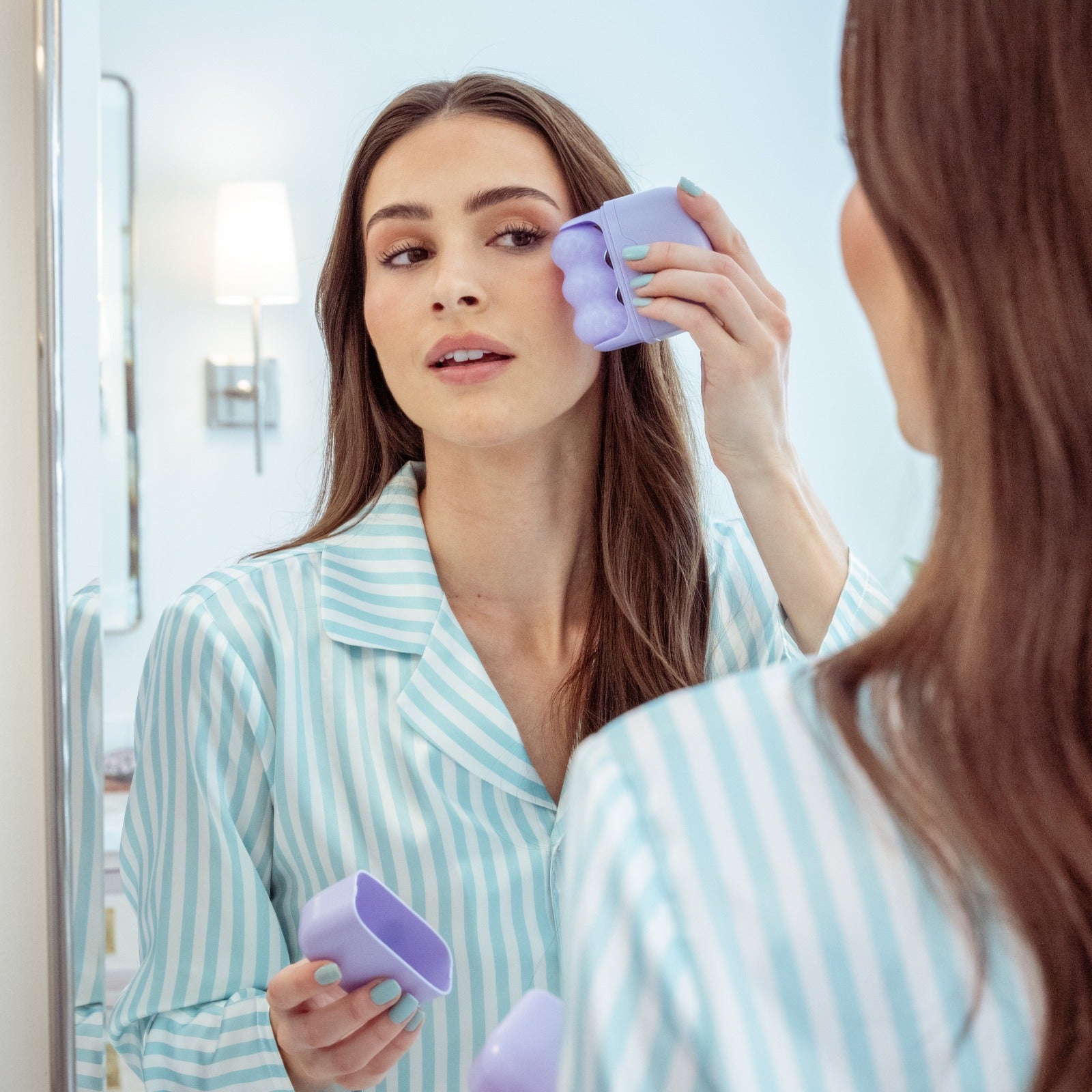 Woman using a lavender three-lobed facial roller on her cheek while looking in a mirror and holding the matching lavender protective cover