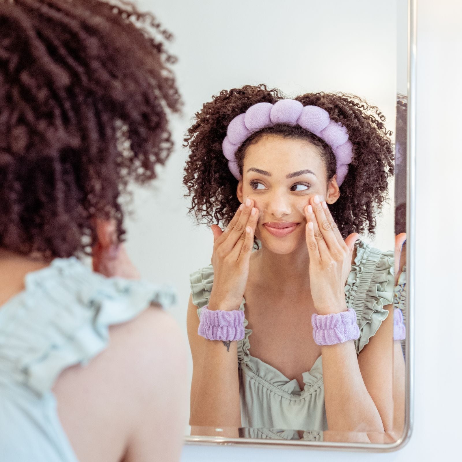 Woman wearing a lavender padded spa headband and matching lavender wristbands while applying skincare to her face in front of a bathroom mirror