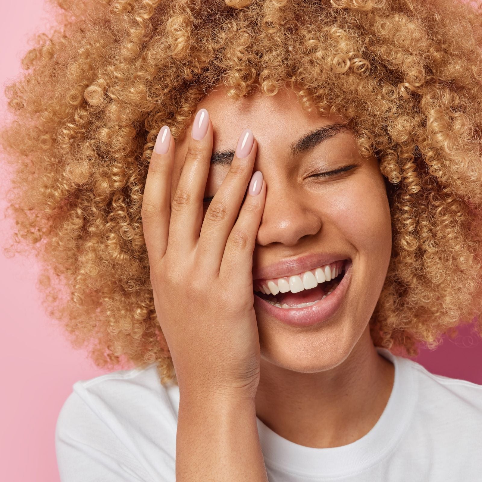 Woman with curly hair showing off a nice, manicured hand smiling against a pink background